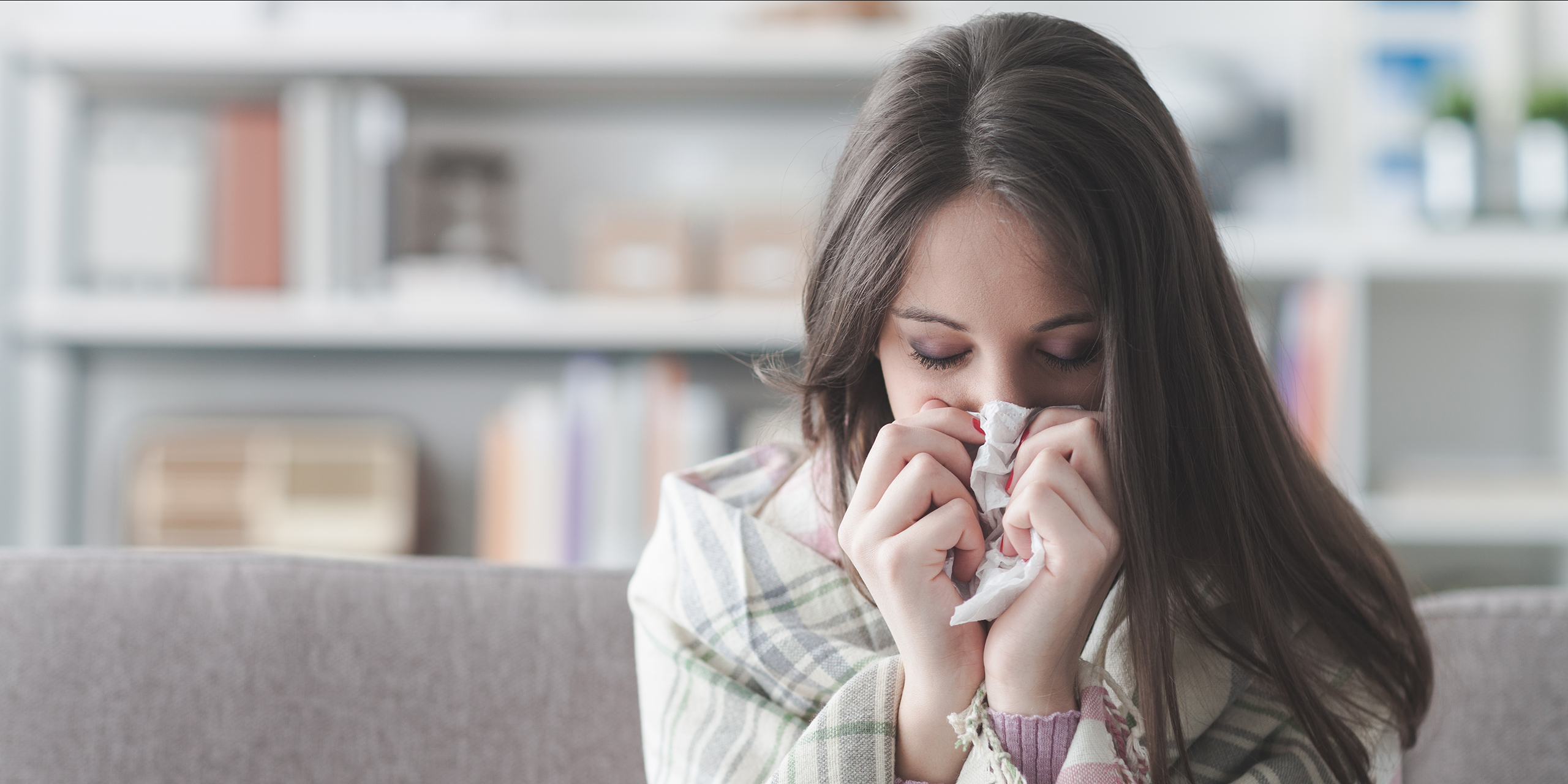 The image depicts a young woman appearing unwell, likely suffering from a cold or flu. She is seated on a grey sofa, partially covered with a plaid blanket in muted tones of green, beige, and white, suggesting she is seeking warmth and comfort. Her head is bowed, and her eyes are closed, conveying a sense of fatigue and discomfort. She is holding a crumpled tissue to her nose, indicating she is blowing her nose, a common symptom of illness. Her long, brown hair is loose and falls around her shoulders, framing her face. The background is softly blurred, showing a bright, modern living room with a white bookshelf filled with books and decorative objects. The bookshelf adds a sense of domesticity and suggests a comfortable, home environment. The overall lighting is soft and natural, contributing to the feeling of vulnerability and illness. The focus is clearly on the woman and her state of being, emphasizing her physical discomfort and need for rest. The image evokes empathy and suggests a moment of quiet suffering within a peaceful home setting. The composition and color palette create a calming yet melancholic mood.